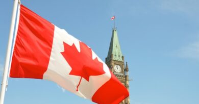 Canadian flag waving in the foreground with the Peace Tower of Parliament Hill in Ottawa against a blue sky