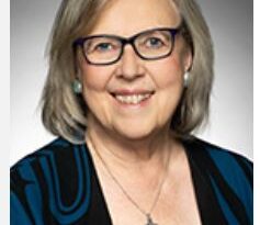 Portrait of a smiling woman with short gray hair and glasses, wearing a blue patterned jacket and a cross necklace.