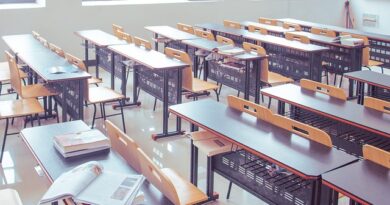 Empty classroom with rows of wooden desks and chairs, papers scattered on a foreground desk and large windows in the background.