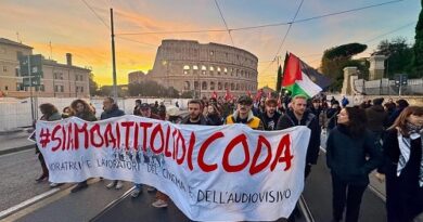 Group of protesters marching on a city street at sunset, carrying a large white banner with bold red text and a Palestinian flag; the Colosseum is visible in the background, with tram lines overhead and bystanders on the sides.