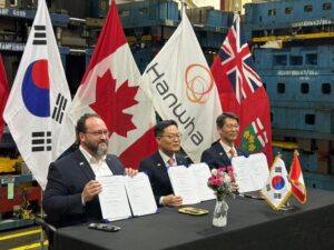 Three men at a signing ceremony hold documents, with national and corporate flags behind them.