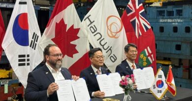Three men at a signing ceremony hold documents, with national and corporate flags behind them.