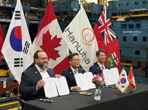 Three men at a signing ceremony hold documents, with national and corporate flags behind them.