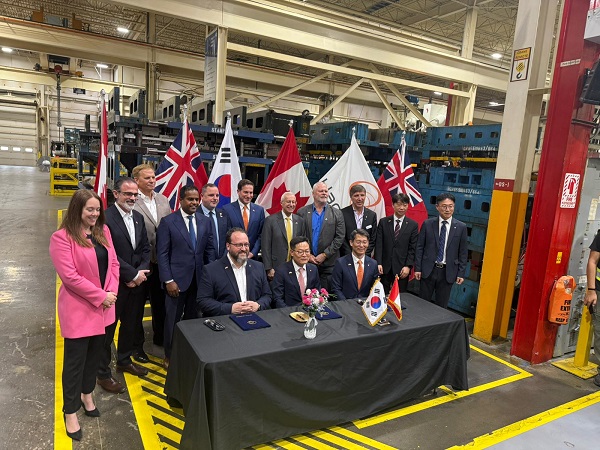Group of officials at a signing ceremony in a factory, with two men seated at a table signing documents and several national flags displayed behind them.