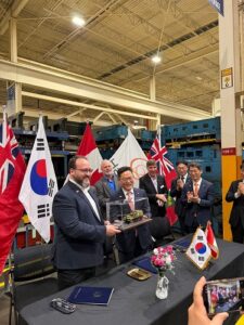 Businessmen in suits at a ceremony around a table with national flags (South Korea, United Kingdom, others) in an industrial showroom, presenting a glass display case with a small model vehicle; several people clap and smile.