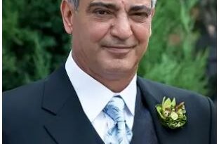 Portrait of a man in a dark suit with a boutonniere, outdoors, facing the camera with a slight smile.
