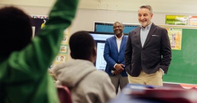Two men in blazers stand at the front of a classroom as a student in the foreground raises his hand to ask a question.