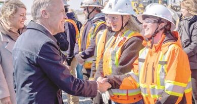 Man in a suit shakes hands with a female construction worker wearing a white helmet and orange safety vest at a busy construction site.