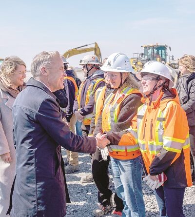 Man in a suit shakes hands with a female construction worker wearing a white helmet and orange safety vest at a busy construction site.
