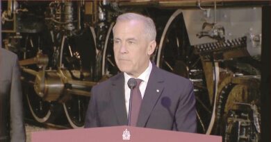 Man in a dark suit and maroon tie speaks at a podium with two microphones, industrial machinery in the background.