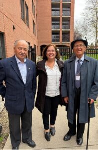 Three adults posing outdoors in front of a brick building: a man in a blue blazer on the left, a woman in a light blouse in the center, and an older man with a hat and cane on the right.