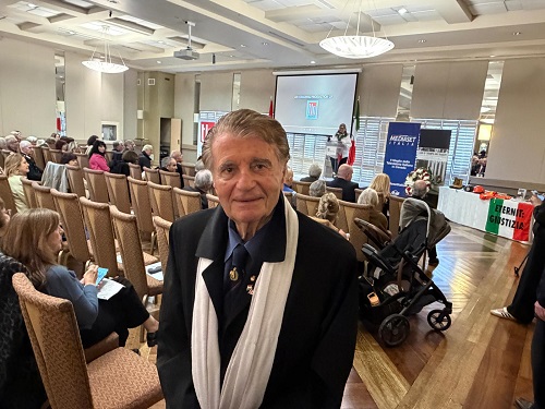 Older man in a dark suit and white scarf posing in a conference hall with rows of chairs and a projector screen behind him.