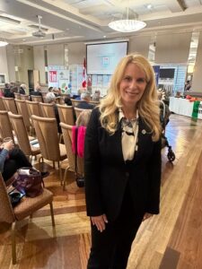 Professional woman in a black suit standing in a conference room with attendees seated and a presentation screen in the background.