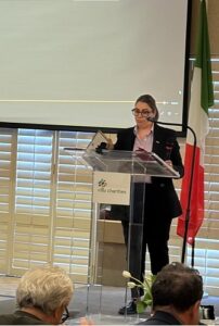 Woman in a blazer giving a speech at a clear podium with a Villa Charities logo; an Italian flag stands behind her.