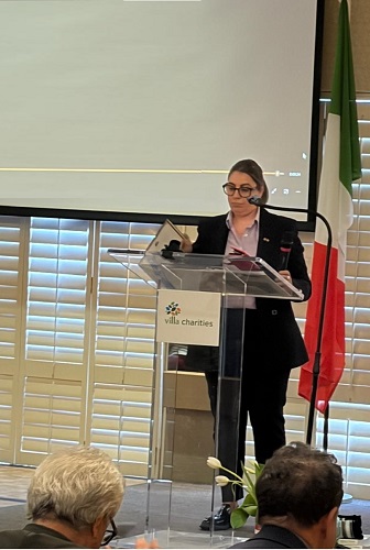 Woman in a blazer giving a speech at a clear podium with a Villa Charities logo; an Italian flag stands behind her.