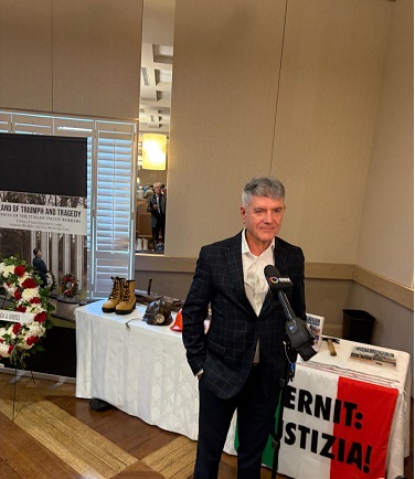 Man in a dark checkered suit speaks into a microphone at a press interview, with a table of shoes and a floral wreath visible in the background.
