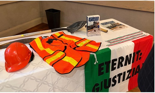 Table display of orange hard hat, safety vests, gloves, and brochures beside a green-white-red banner reading 'ETERNIT: GIUSTIZIA' on the tablecloth.