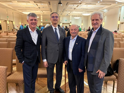 Four men in business suits posing for a photo in a conference hall; one older man is holding a cane.