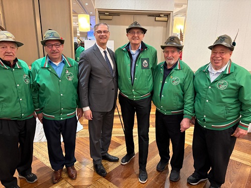 Six men pose together in a hotel lobby; five wear green jackets with patches and hats, one in a suit.