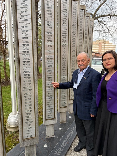 An elderly man in a dark blue suit and a woman in a purple blazer stand beside a tall memorial wall covered with vertical name plaques.
