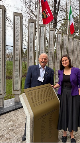 Elderly man in a dark suit and a woman in a purple blazer pose at a monument with tall engraved stone tablets and flags in the background.