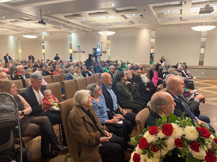 Large audience seated in a conference hall during an event, with a floral arrangement in the foreground and a camera setup near the back.