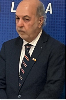 Older man in a dark suit and striped tie standing at a podium with a blue backdrop, wearing a lapel pin.