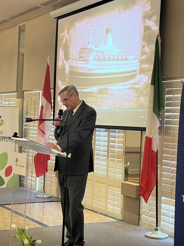 Man in a suit speaking at a podium with a microphone in a conference room, flags behind him and a ship image projected on a screen.