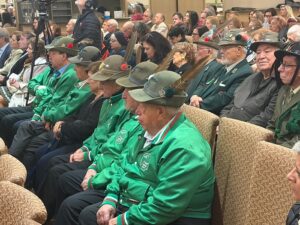 Audience of veterans in green jackets and hats sits in a conference hall, watching a ceremony.