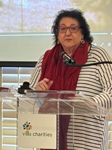 Woman with glasses and a red scarf speaks at a transparent lectern bearing the Villa Charities logo.