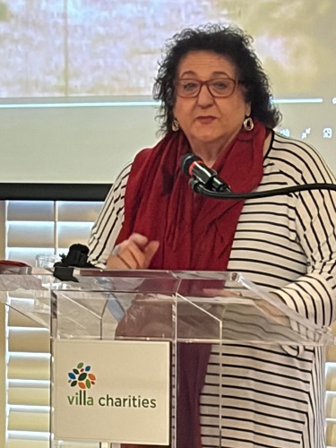 Woman with glasses and a red scarf speaks at a transparent lectern bearing the Villa Charities logo.