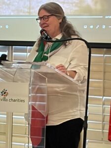 Woman with glasses and gray hair delivers a talk at a clear podium, green scarf and Villa Charities logo visible.