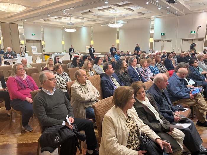 Audience seated in a large conference hall, listening to a presentation at the front of the room.