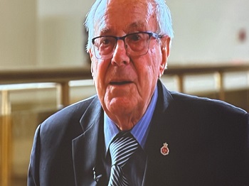 Older man with gray hair and glasses, wearing a dark suit and striped tie, speaking.