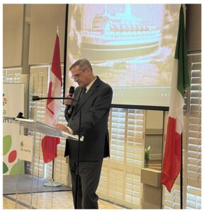 Speaker at a podium delivering a formal talk, with a large screen displaying a ship image behind him and flags of Canada and Italy nearby in a conference room.