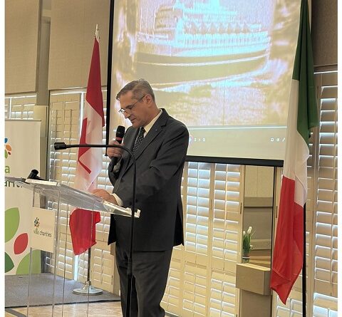 Speaker at a podium delivering a formal talk, with a large screen displaying a ship image behind him and flags of Canada and Italy nearby in a conference room.