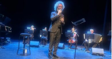 Male singer in a dark suit performs center stage with a microphone, backed by a small ensemble of violinists, a cellist, and an accordionist under blue stage lighting.