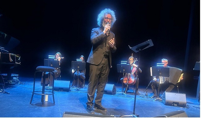 Male vocalist with microphone center stage, blue-lit concert with a string ensemble behind him on risers