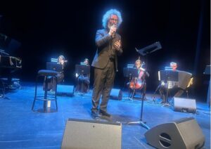 Male singer with curly hair and a beard in a black suit performing on a dark stage, with a small ensemble behind him.