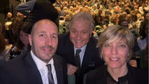 Three professionally dressed attendees pose for a photo in a crowded auditorium.