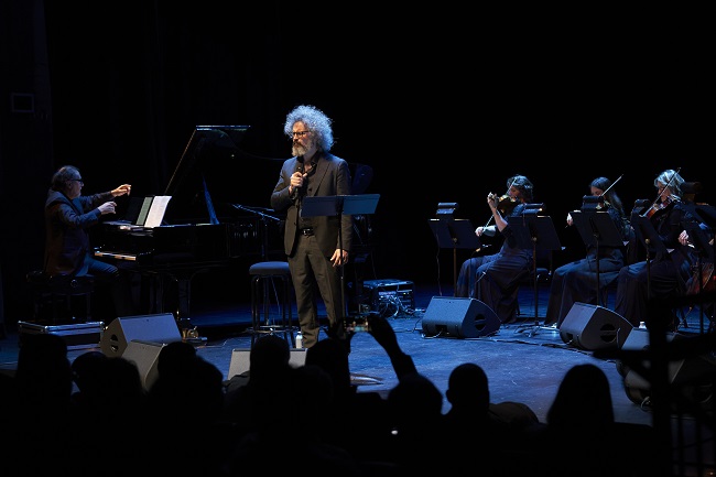 Male singer with curly hair performs center stage with microphone, pianist on left and string ensemble on right during a live concert.