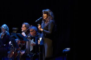 Female singer at center stage with a microphone, performing with a small band under blue stage lighting (violin/accordion players visible in the background).
