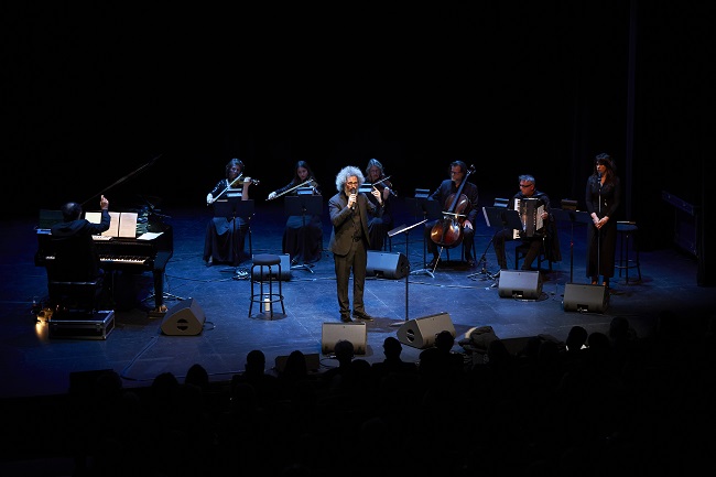 Male vocalist in a suit performs center stage with a small ensemble, piano on the left, lit by blue spotlight in a dark theater; audience silhouettes in front.