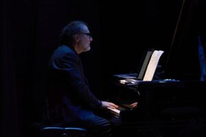 Pianist playing a grand piano on a dark stage, illuminated by a blue light with sheet music in front of him at eye level.