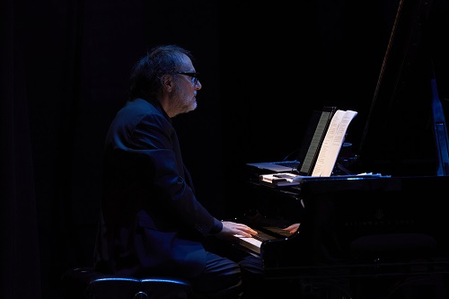 Pianist playing a grand piano on a dark stage, illuminated by a blue light with sheet music in front of him at eye level.