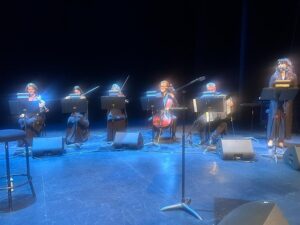 A row of six musicians seated on stage, playing string instruments (violin and cello) with a vocalist at a lectern on the right under blue stage lighting