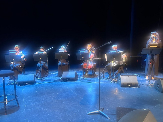 Six musicians seated on a stage with music stands, playing violins and a cello, while a woman at the far right sings into a microphone.