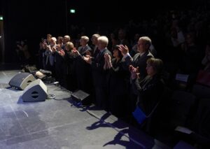 Group of formally dressed people standing at the stage edge, clapping during a live event, with a dark audience in the background.