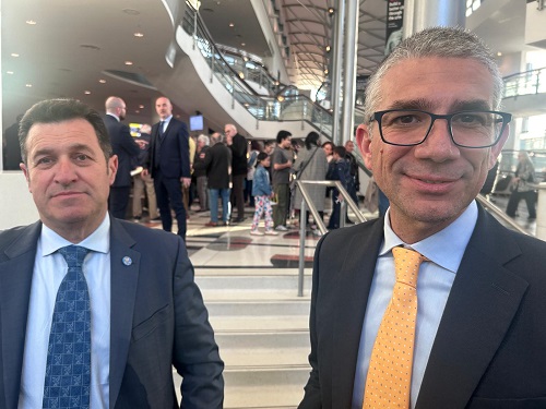 Two men in suits pose for a photo in a modern atrium with stairs and a crowd in the background; one wears a blue patterned tie and the other an orange tie.