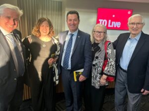 Group of five professionally dressed people posing together at an event, with a red 'TO Live' screen in the background.